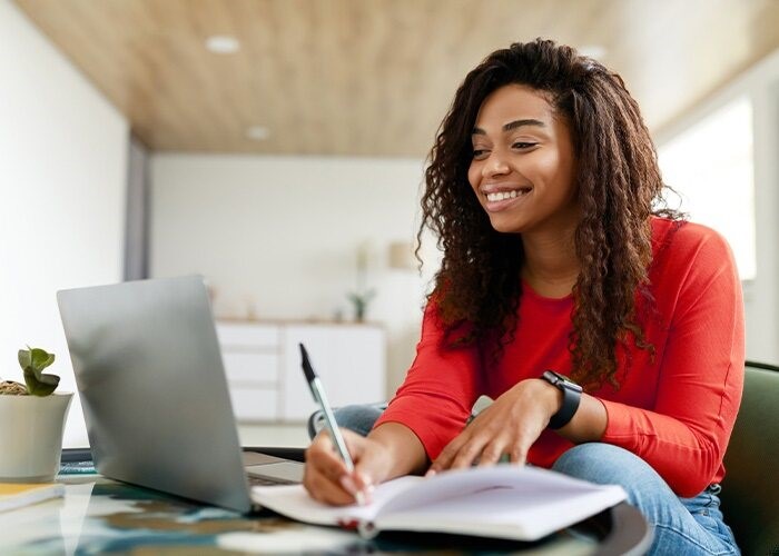 A young woman sits at a table with a laptop and notebook while she takes notes by hand.
